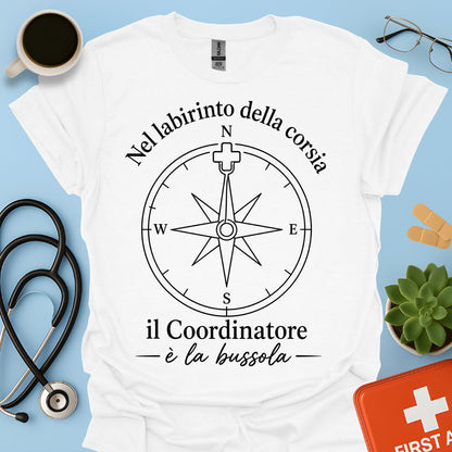 Maglietta t-shirt bianca a maniche corte da infermiere con grafica nera di una bussola e la frase: "Nel labirinto della corsia il Coordinatore è la bussola" con ago a forma di croce medica.